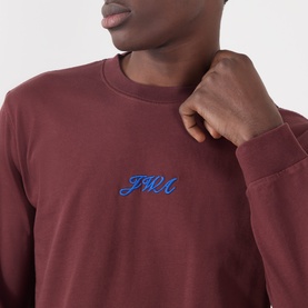 Model wearing JW Anderson burgundy long-sleeve T-shirt with blue cursive logo embroidery at chest, in cotton fabric.