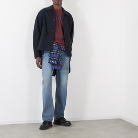 Model wearing JW Anderson burgundy T-shirt styled with dark overshirt, blue plaid shirt tied at waist, and mid-wash straight jeans.
