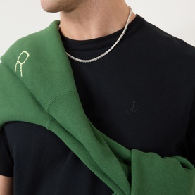Model wearing JW Anderson Anchor Logo T-Shirt in black with green hoodie draped over shoulder and silver chain necklace.