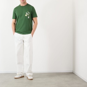 Model wearing JW Anderson Scattered Logo T-Shirt in green with chest graphic, styled with white straight-leg trousers and white trainers.