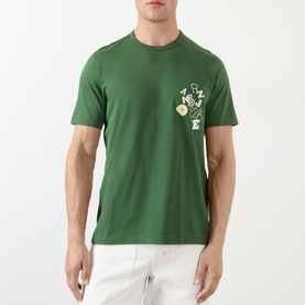 Model wearing JW Anderson Scattered Logo T-Shirt in green with scattered lettering graphic on chest, styled with white trousers.