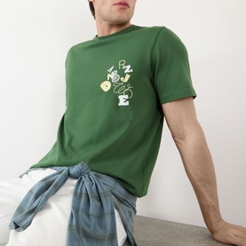 Model wearing JW Anderson Scattered Logo T-Shirt in green with scattered lettering graphic on chest, styled with blue jeans tied at waist.
