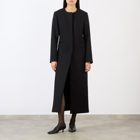 Model wearing Loulou Studio black wool blend long coat with collarless neckline and front slit, styled with black heels and dress.