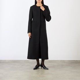 Model wearing Loulou Studio black wool blend longline coat with collarless neckline and front pockets, styled with black heels and tights.