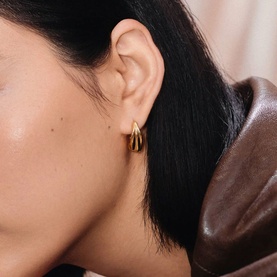 Model with Missoma gold chunky hoop earrings, shown as a pair on ear, with warm-toned draped fabric in background