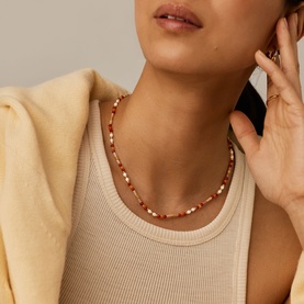 Model with Missoma gold red beaded necklace featuring mixed red and gold-tone beads, worn with cream ribbed top and yellow jacket