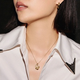 Model with Missoma gold pendant necklace featuring embossed heart locket on delicate chain, styled with gold hoop earrings and white shirt collar
