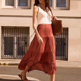Model wearing a rust tiered maxi skirt, styled with a white vest top, carrying a woven basket bag, and brown loafers