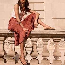 Model wearing rust midi skirt and white vest top, seated on stone balustrade, styled with tan strappy flat sandals