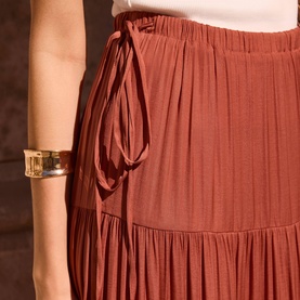 Model wearing a rust-coloured pleated tiered midi skirt with elasticated waist and side tie detail, styled with a gold cuff bracelet