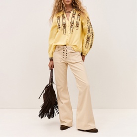 Model wearing Bash yellow embroidered blouse with wide-leg cream trousers, carrying brown fringed suede bag