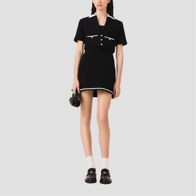 Model wearing black mini dress with white contrast trim, short sleeves, and button detail, styled with white socks and black loafers