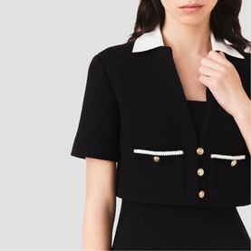 Model wearing black short-sleeve tweed jacket with white contrast trim, pearl-edged pockets, and gold buttons, styled over a black dress.