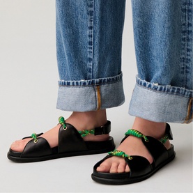 Model wearing black flat sandals with green rope lace detailing and ankle strap, styled with cuffed blue denim jeans