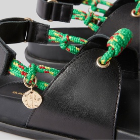 Black leather sandal detail showing green and red rope strap with gold charm and eyelet hardware
