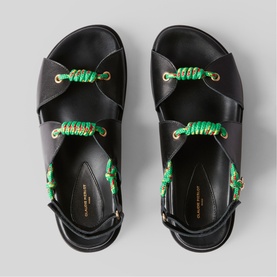 Black leather crossover sandals with green braided rope lace detailing and gold hardware, shot from above on grey background