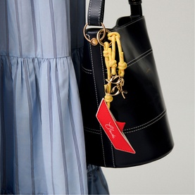 Model carrying black leather bucket bag with gold-tone hardware, yellow rope keyring charm, and red branded tag detail, styled with blue striped