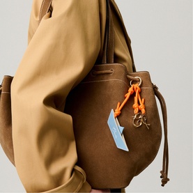 Model carrying a brown suede bucket bag with orange drawstring hardware and light blue charm keyring detail