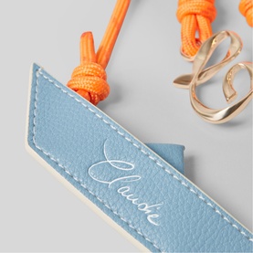 Light blue leather Laude branded tag with orange cord and gold hook clasp detail, close-up shot