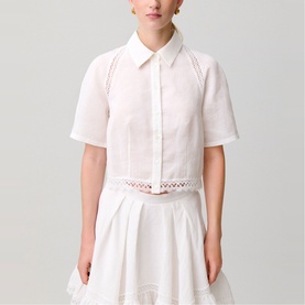 Model wearing white cropped short-sleeve shirt with lace trim waistband and subtle pink stitch detail, styled with white pleated skirt