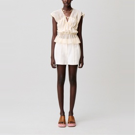 Model wearing cream ruffled sleeveless top with tie neck and matching tiered mini skirt, styled with red patterned flat shoes