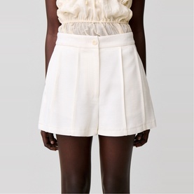 Model wearing white tailored shorts with pleated front and button detail, styled with a cream ruffled top