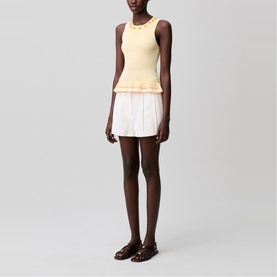 Model wearing yellow sleeveless peplum knit top with white mini skirt and brown sandals, full-length view