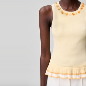 Model wearing a pale yellow sleeveless ribbed top with orange floral embellishment at neckline, styled with a pleated skirt