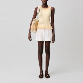 Model wearing yellow sleeveless knit top with ruffle hem and white tailored shorts, styled with a woven tote bag and brown sandals