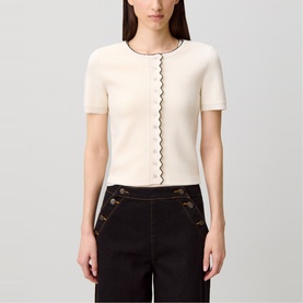 Model wearing cream short-sleeve knit top with black zigzag trim detail, styled with dark tailored trousers with gold button embellishment.