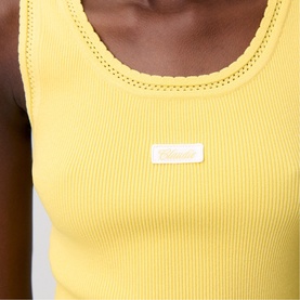 Model wearing yellow ribbed sleeveless vest top with small logo badge detail at chest