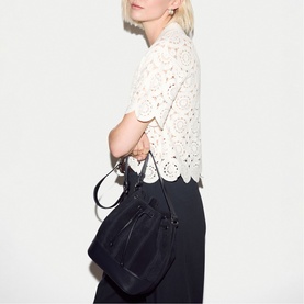 Model carrying Jigsaw navy bucket bag with drawstring closure and tassel detail, styled with white broderie top and black trousers.