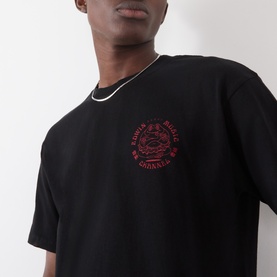 Model wearing Edwin black crew neck T-shirt with red circular graphic logo print on chest, styled with a silver chain necklace.