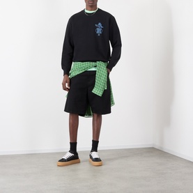 Model wearing Edwin black cotton sweatshirt with graphic print, styled with black shorts and green check shirt tied at waist.