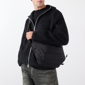Model carrying Lemaire Medium Soft Game Shoulder Bag in dark chocolate, worn crossbody over black textured zip-up jacket with grey jeans.