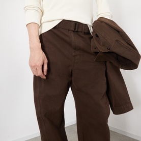 Model wearing Lemaire Belted Casual Trousers in brown with matching belt, styled with a cream knit top and brown jacket draped over arm.