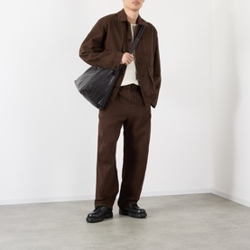 Model wearing Lemaire Belted Casual Trousers in brown, styled with matching brown jacket, cream top, and black chunky loafers. Model carries a black