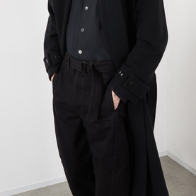 Model wearing Lemaire black cotton casual trousers with wide leg fit, styled with matching black shirt and longline jacket.