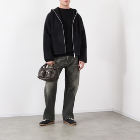 Model wearing Lemaire black wool crew neck, styled with dark wash jeans, zip-up hoodie, and loafers, carrying dark holdall bag.