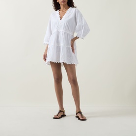 Model wearing Juliet Dunn white cotton mini dress with V-neckline, balloon sleeves, tiered skirt and broderie anglaise hem trim, styled with tan