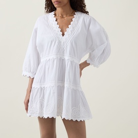 Model wearing white cotton mini dress with V-neckline, puff sleeves, tiered skirt and scalloped broderie anglaise trim throughout.