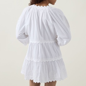 Model wearing Juliet Dunn white cotton mini dress, back view, featuring puff sleeves, scalloped trim, broderie anglaise tiers and belted waist.