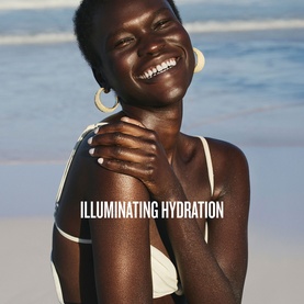 Model with Sol De Janeiro Illuminating Hydration skincare set, smiling on a beach, wearing white swimwear and gold hoop earrings.