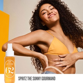 Model with Sol De Janeiro Hydrating Jet Set Cheirosa 62 spray bottle, wearing yellow bikini top, smiling against orange background.