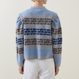 Model wearing Urban Aran grey merino wool cardigan, rear view, with blue and charcoal geometric brick-pattern knit, styled with white trousers