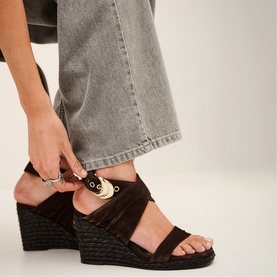 Model wearing Bash brown suede wedge sandals with gold buckle detail and cross-strap, styled with grey wide-leg jeans