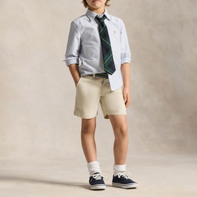 Model wearing Ralph Lauren light blue stripe children's shirt with green tartan tie, styled with beige shorts and navy canvas shoes.