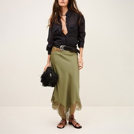 Model wearing Bash green satin midi skirt with lace hem, styled with black shirt, western belt, and strappy sandals, carrying black feather bag