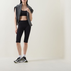 Model wearing Tala Shadow Black cycling leggings with a black sports bra and grey zip-up vest, styled with white socks and chunky trainers.