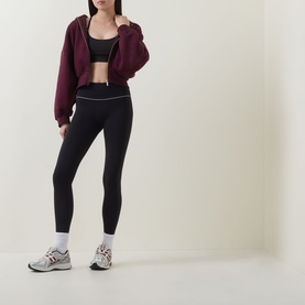 Model wearing Tala Shadow Black leggings with white contrast waistband, styled with a cropped burgundy hoodie, black sports bra and grey trainers.